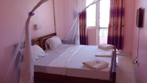 A single bed with a canopy, a bedside table, a wall-mounted air conditioner, and a window with curtains. - Checkpoint Inn (Mombasa)
