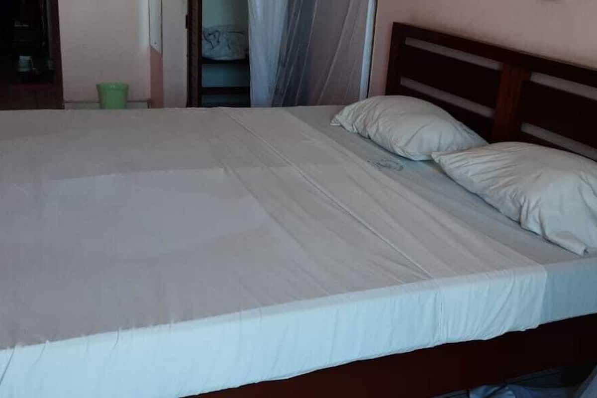 Un lit avec un matelas et des oreillers blancs, une tête de lit en bois et un seau vert posé sur le sol.