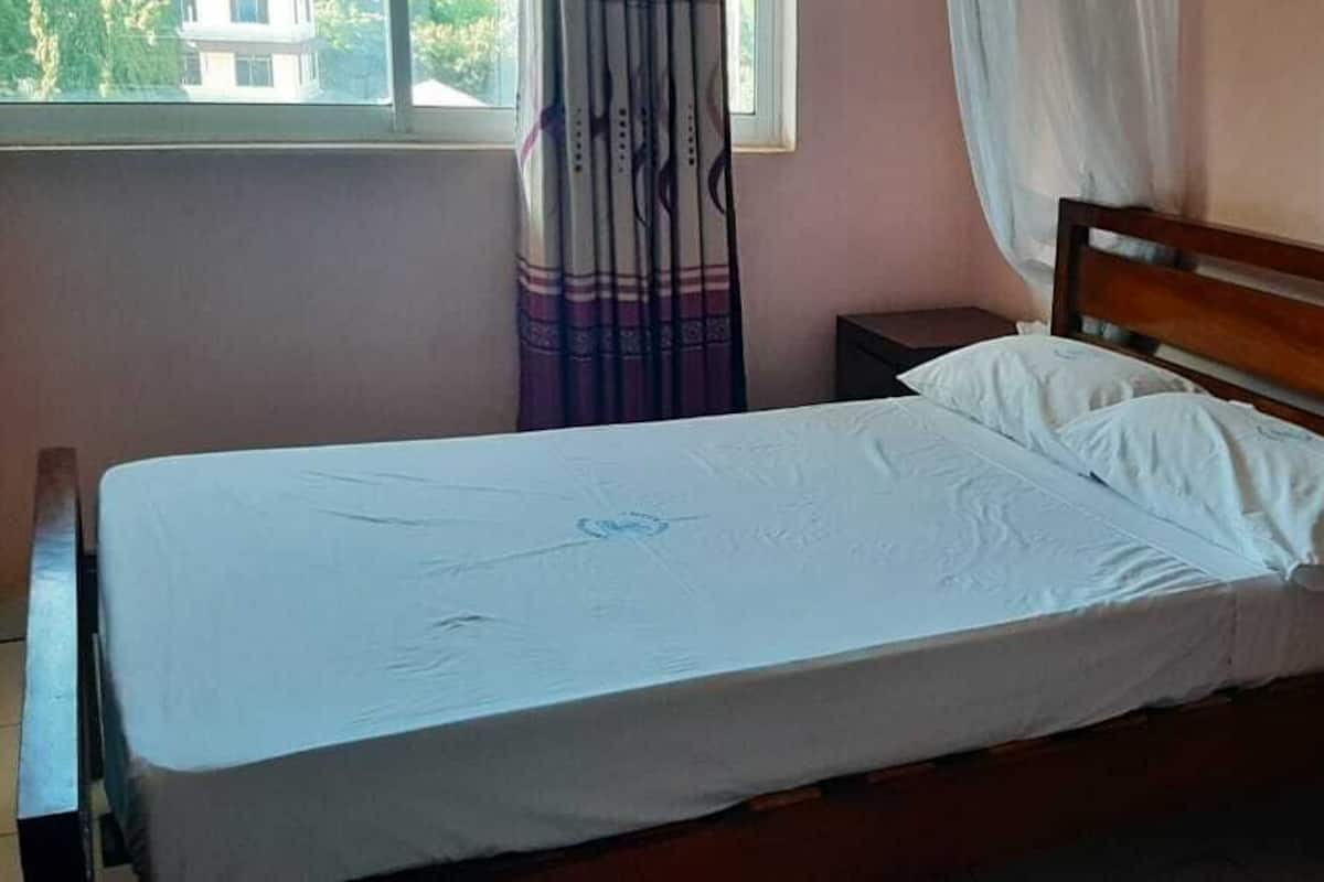 Un lit simple avec une tête de lit en bois, une table de chevet, une fenêtre avec des rideaux et une salle de bain visible à travers un miroir.