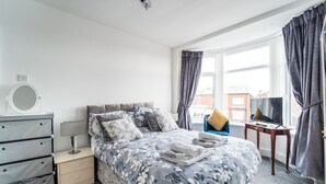 Une chambre à coucher avec un lit, une commode, une télévision et une fenêtre avec des rideaux.