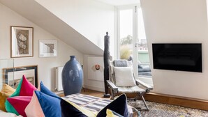 A modern living room with a large window, a flat-screen TV, and a variety of colorful pillows. - Unique 2bed Apartment, Portobello Rd (London)