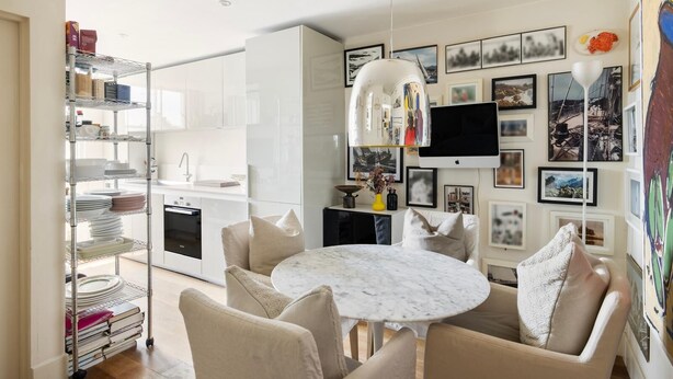 A modern kitchen with a dining area, featuring a marble table, beige chairs, and a glass shelving unit with dishes. - Unique 2bed Apartment, Portobello Rd (London)