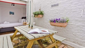 A cozy room with a white brick wall, a bed with white bedding, a wooden table with an open book, and hanging planters with greenery.