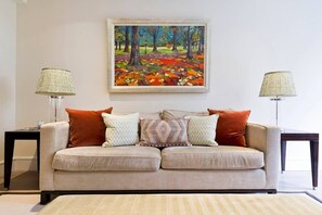 A living room with a beige sofa, patterned cushions, and a painting of a forest scene.