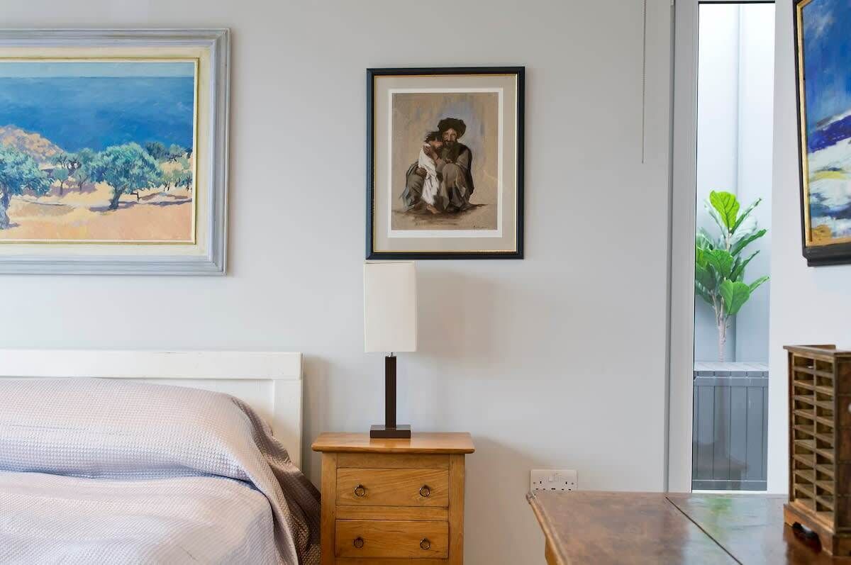 A bedroom with a bed, a nightstand with a lamp, a framed painting, and a view of a balcony with a plant.