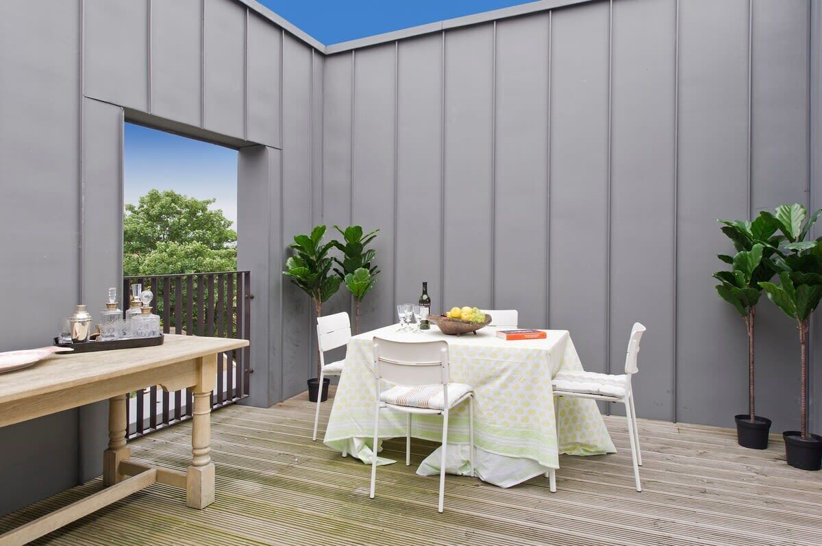 An outdoor patio with a wooden table and chairs, a small garden area with potted plants, and a view of trees through a large window.