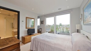 A bedroom with a large bed, a TV on a wooden stand, a balcony with a view, and a sliding glass door to the bathroom. - Fabulous 2-bed Penthouse, Wimbledon (London)