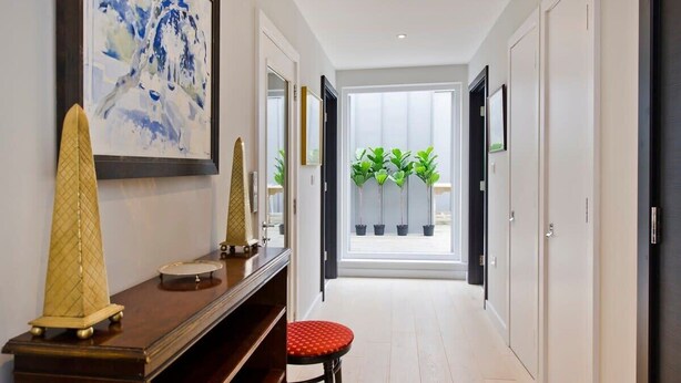 A hallway with a wooden console table, a red stool, a framed abstract painting, and a view of a balcony with potted plants. - Fabulous 2-bed Penthouse, Wimbledon (London)