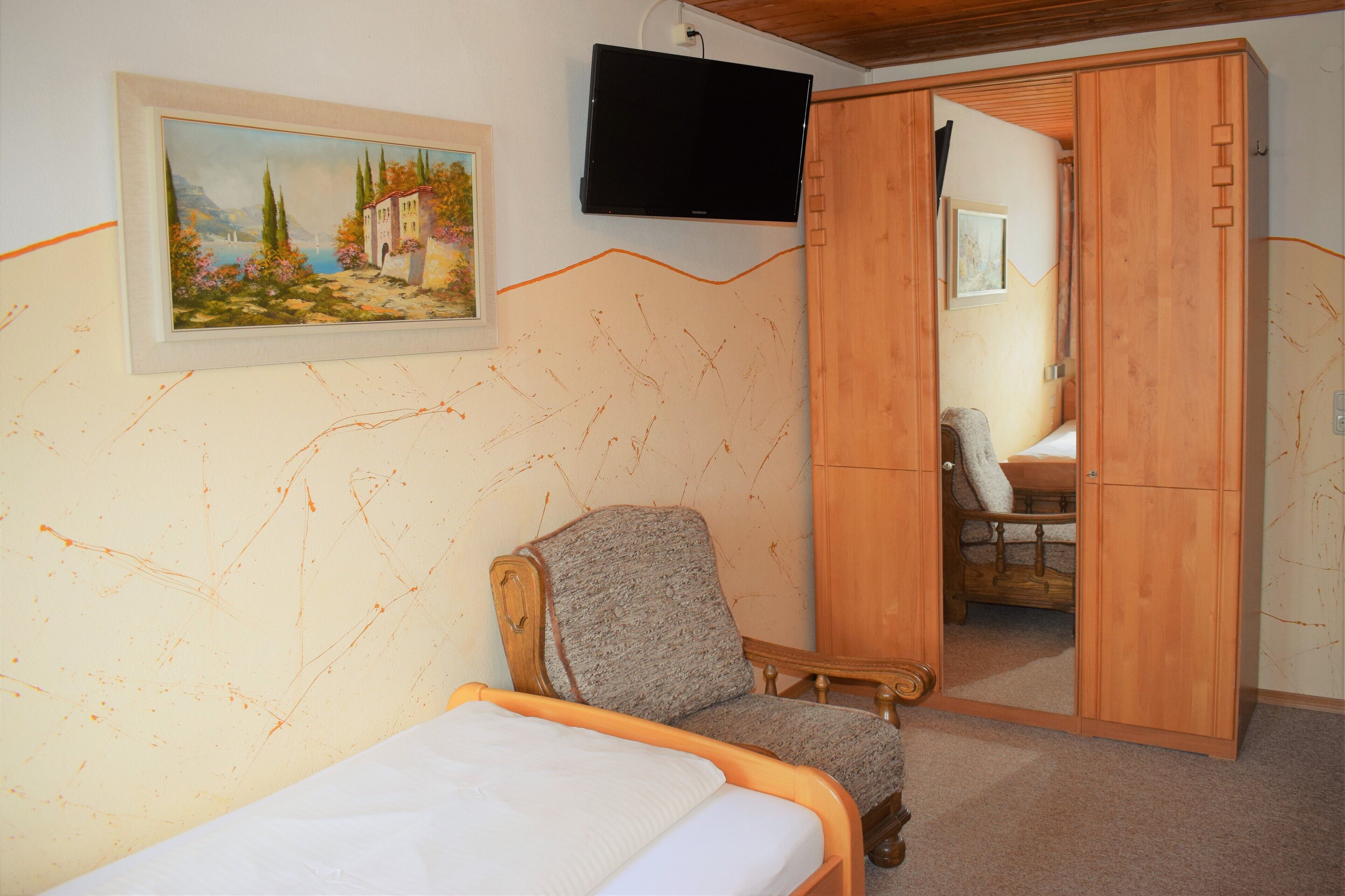 Un dormitorio con cama, una silla, televisión montada en la pared y un cuadro.
