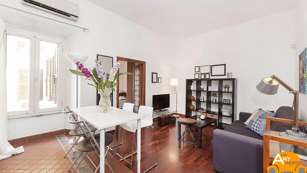 Una sala moderna con una mesa de comedor blanca, un sofá y un televisor. Hay una estantería con objetos decorativos y una lámpara.