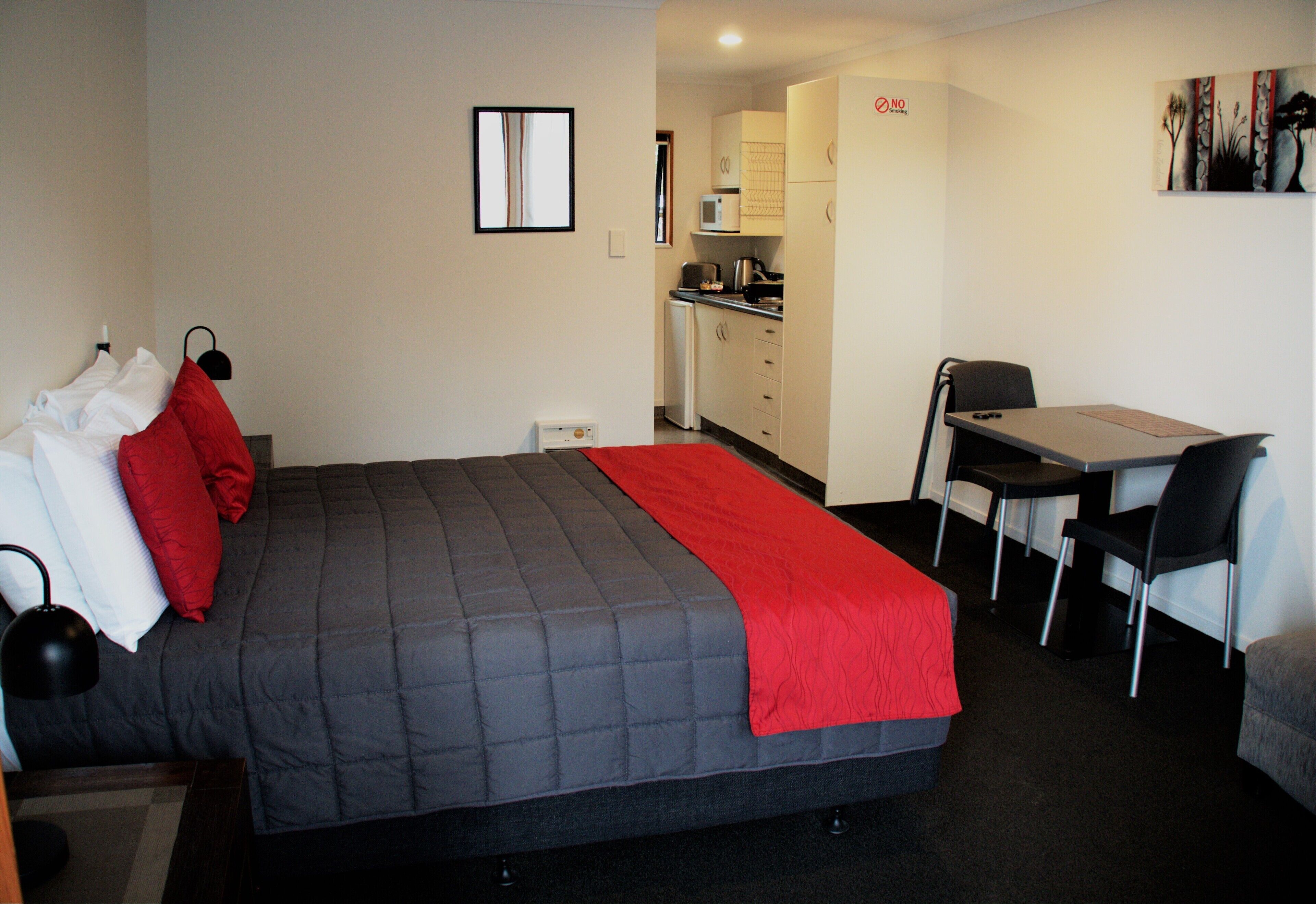 A hotel room with a bed, a desk, a chair, a kitchenette, and a door with a 'NO' sign.