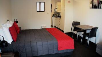 A hotel room with a bed, a desk, a chair, a kitchenette, and a door with a 'NO' sign.