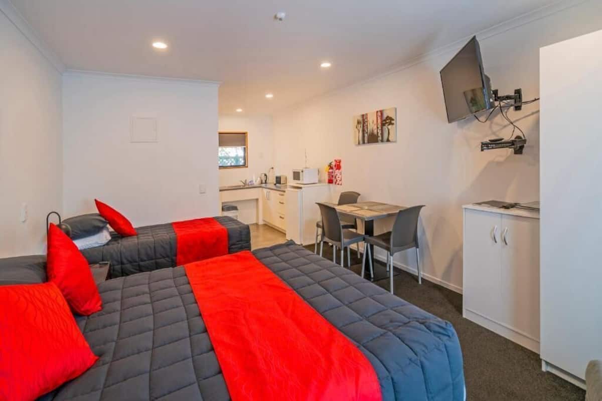A compact hotel room with two beds, a kitchenette, a dining area, and a TV mounted on the wall.