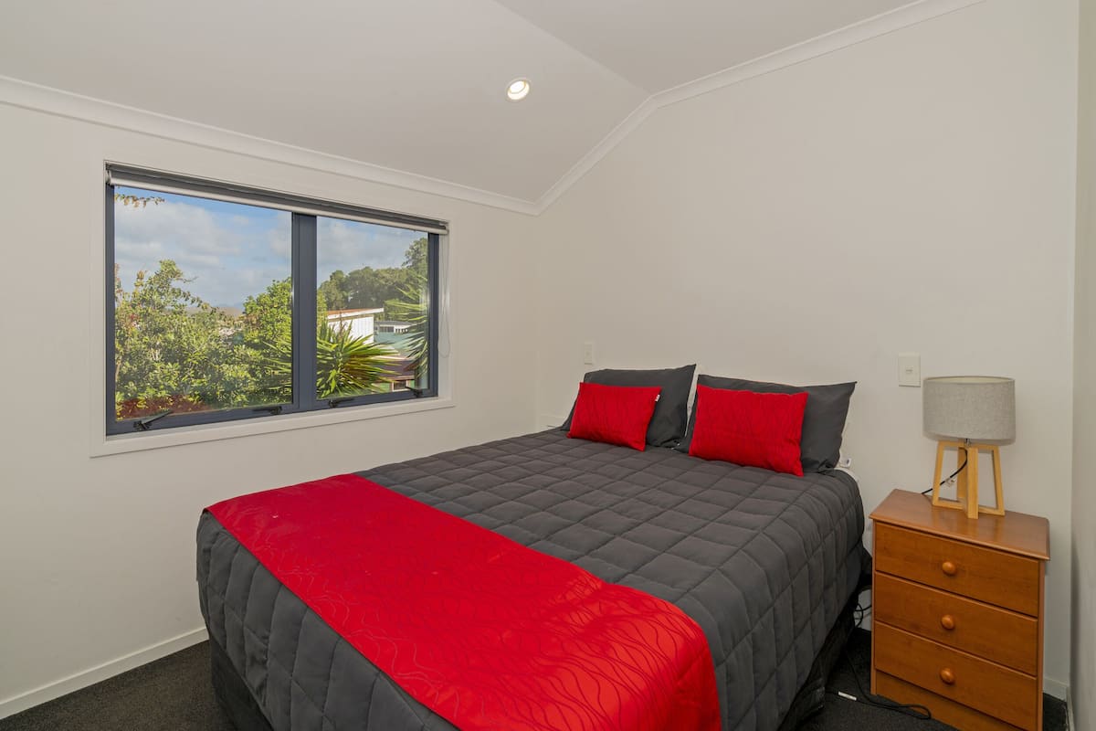 A bedroom with a large bed, a nightstand, a lamp, and a window with a view of greenery.