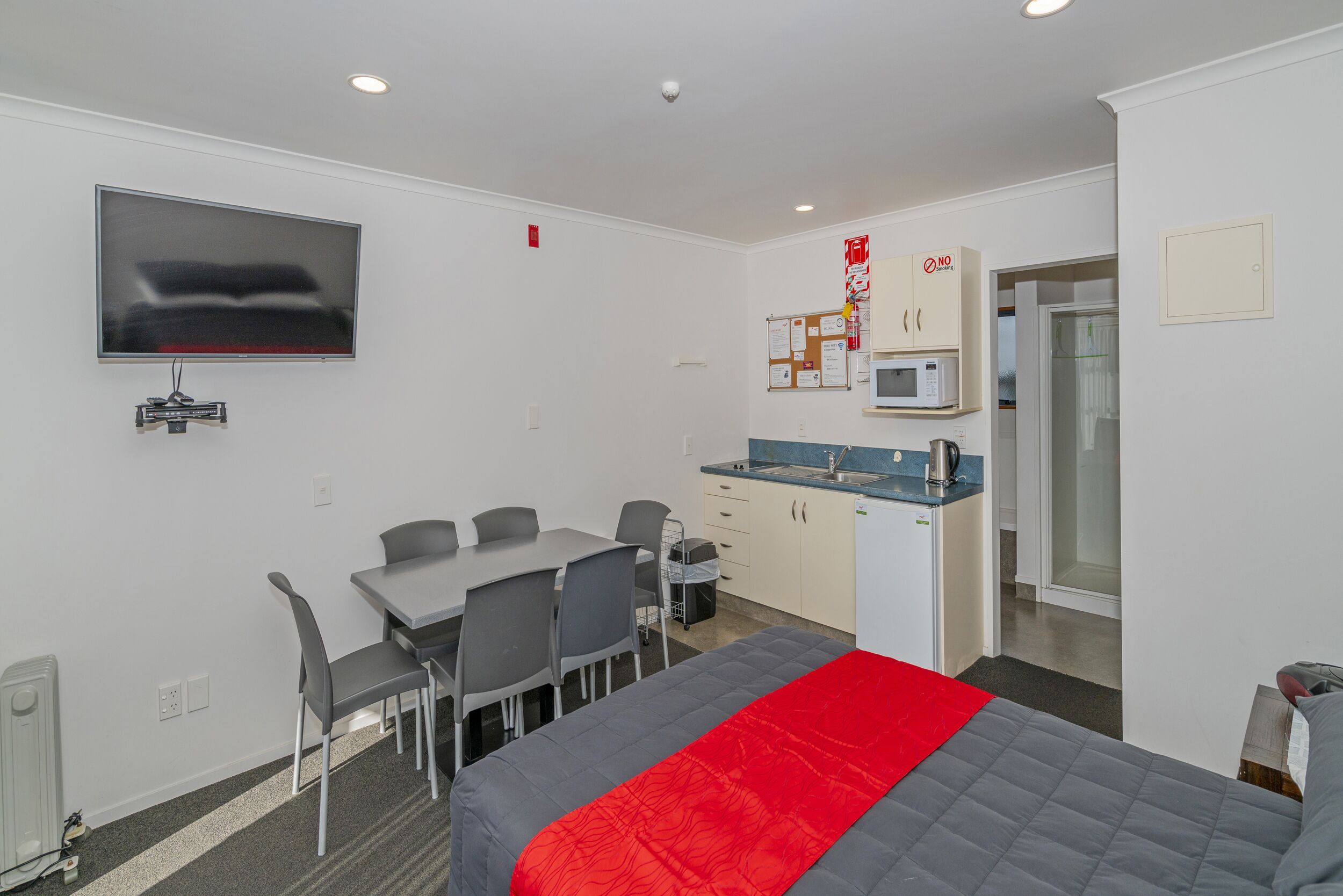 A compact hotel room with a bed, a desk, chairs, a kitchenette, and a TV.