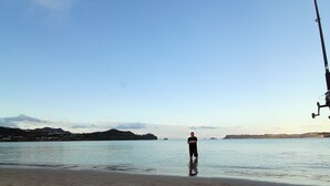Una playa con una persona de pie en aguas poco profundas, una caña de pescar y colinas al fondo.