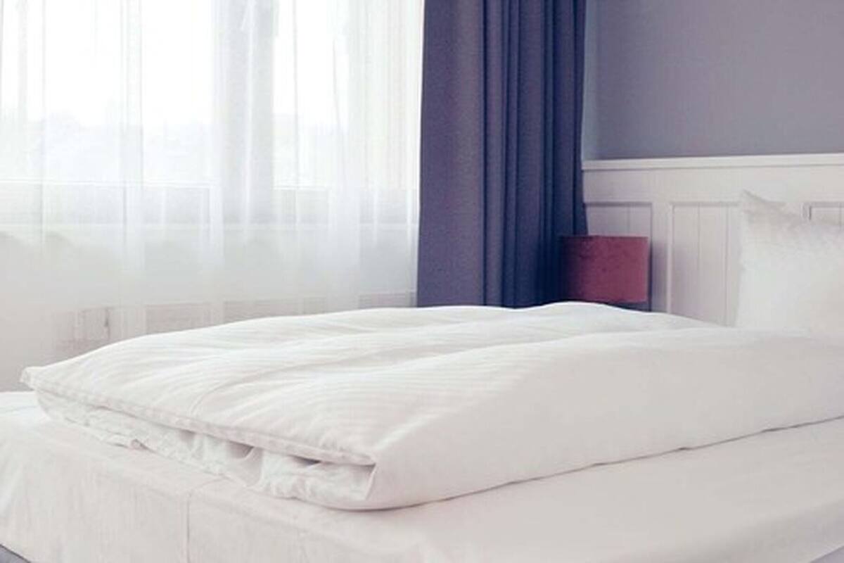 A neatly made bed with white bedding in a room with a window, a blue curtain, and a white radiator.