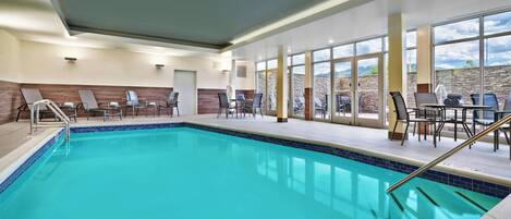 An indoor swimming pool with clear blue water, surrounded by lounge chairs and tables.