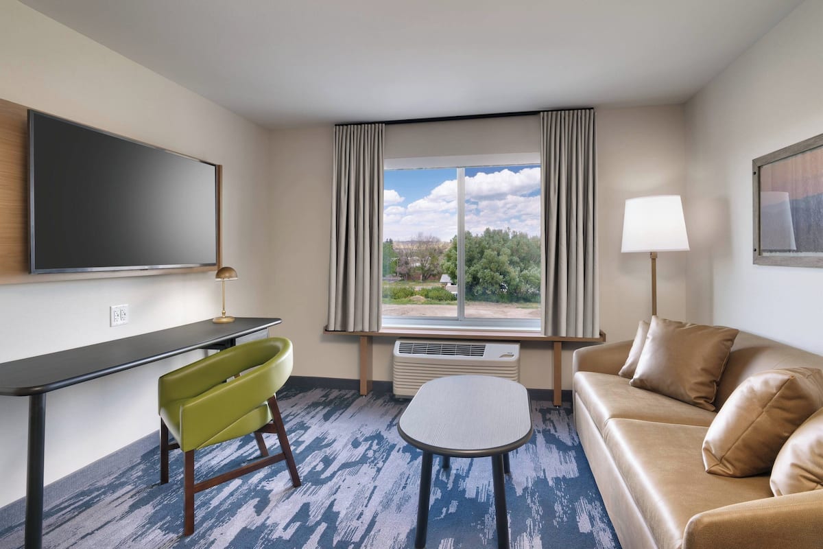 Een hotelkamer met een flatscreen-tv, een groene stoel, een beige bank, een klein tafeltje en een raam met gordijnen.