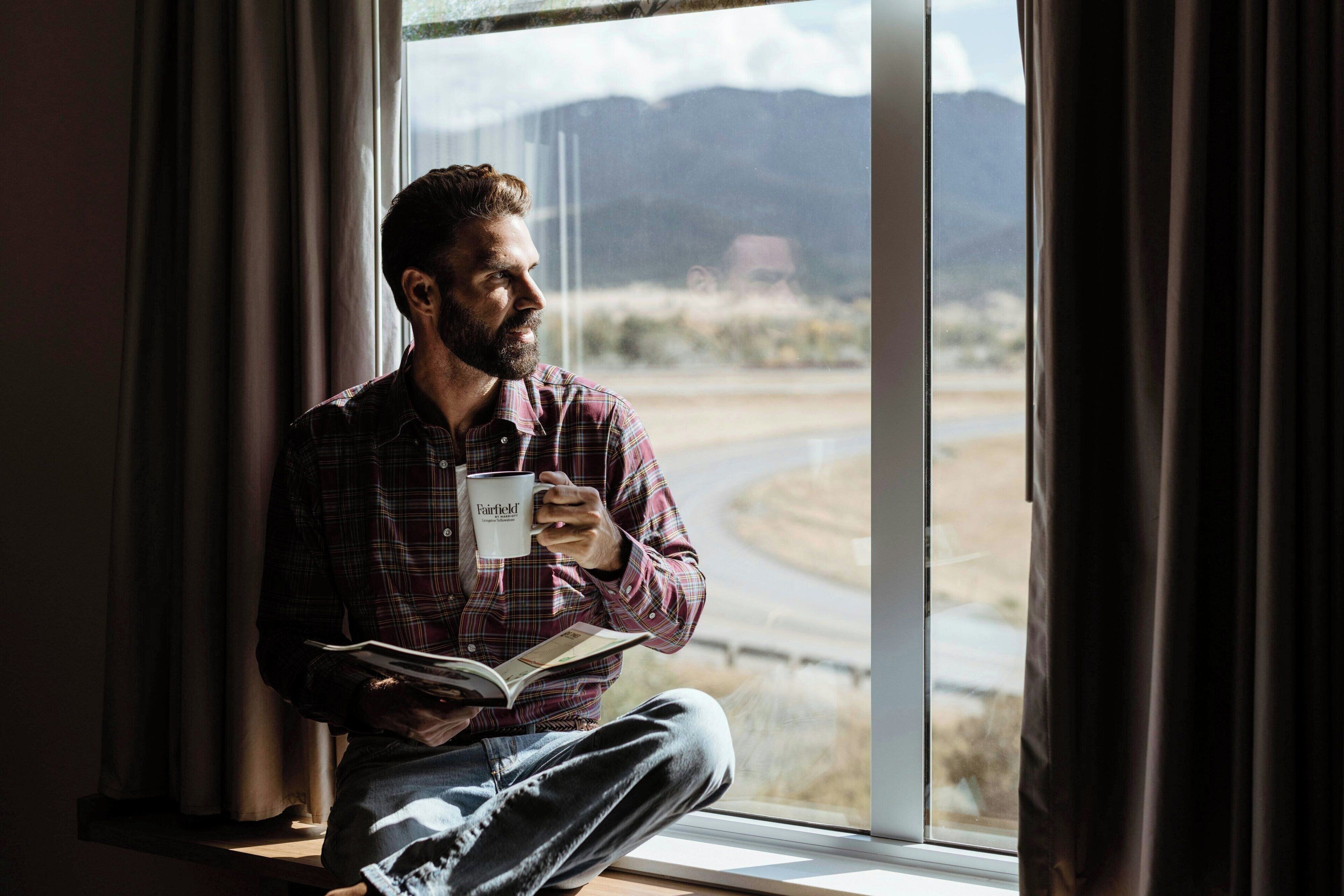 Une personne assise près d’une fenêtre, en train de lire un livre et tenant une tasse.