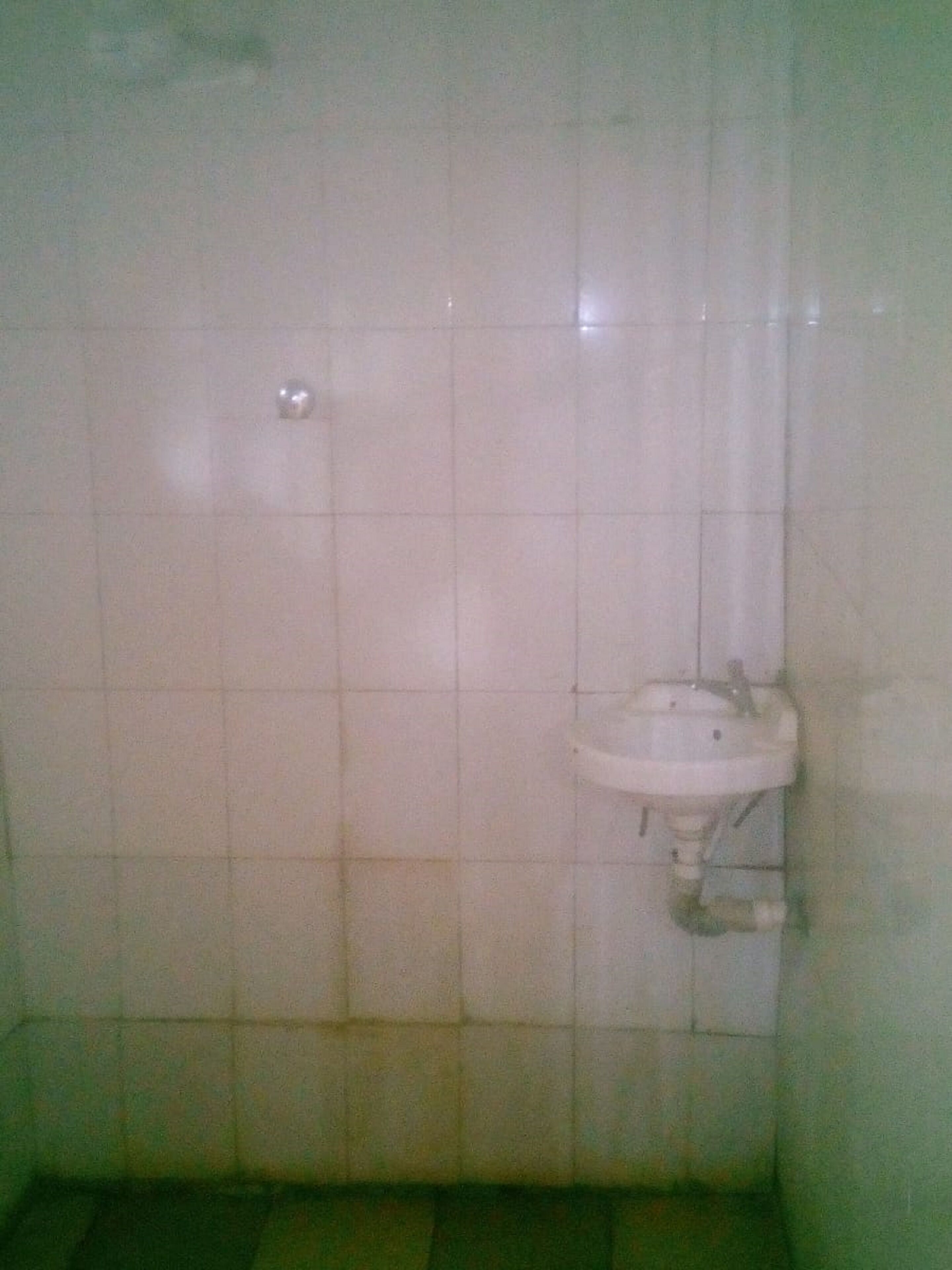 A small, dimly lit bathroom with a single white sink and a mirror. The walls are tiled, and the floor appears to be made of greenish tiles.