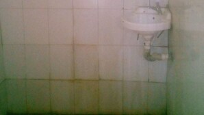 Un baño pequeño y tenuemente iluminado, con un lavamanos blanco y un espejo. Las paredes están revestidas de azulejos, y el suelo parece estar hecho de baldosas verdosas.