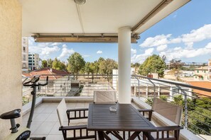 Ein Balkon mit Tisch und Stühlen, einem feststehenden Ergometer und Blick auf die umliegenden Gebäude und Bäume.