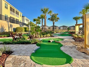 A miniature golf course with a green putting area, palm trees, and a building in the background.