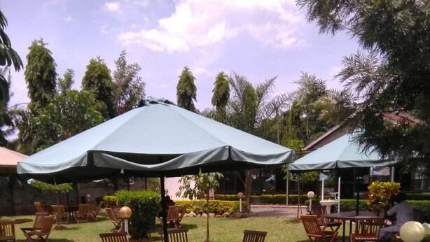 An outdoor seating area with wooden tables and chairs, covered by large umbrellas, surrounded by greenery and trees. - EL Paso Gardens Kisumu (Kisumu)