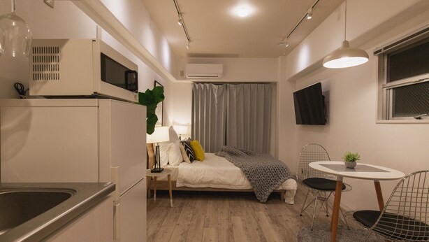 A compact room with a kitchenette, a bed with a grey blanket, a small dining table with chairs, and a TV mounted on the wall. - Akebono 5-A (Naha)