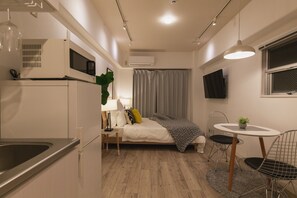 A compact room with a kitchenette, a bed with a grey blanket, a small dining table with chairs, and a TV mounted on the wall. - Akebono 5-A (Naha)