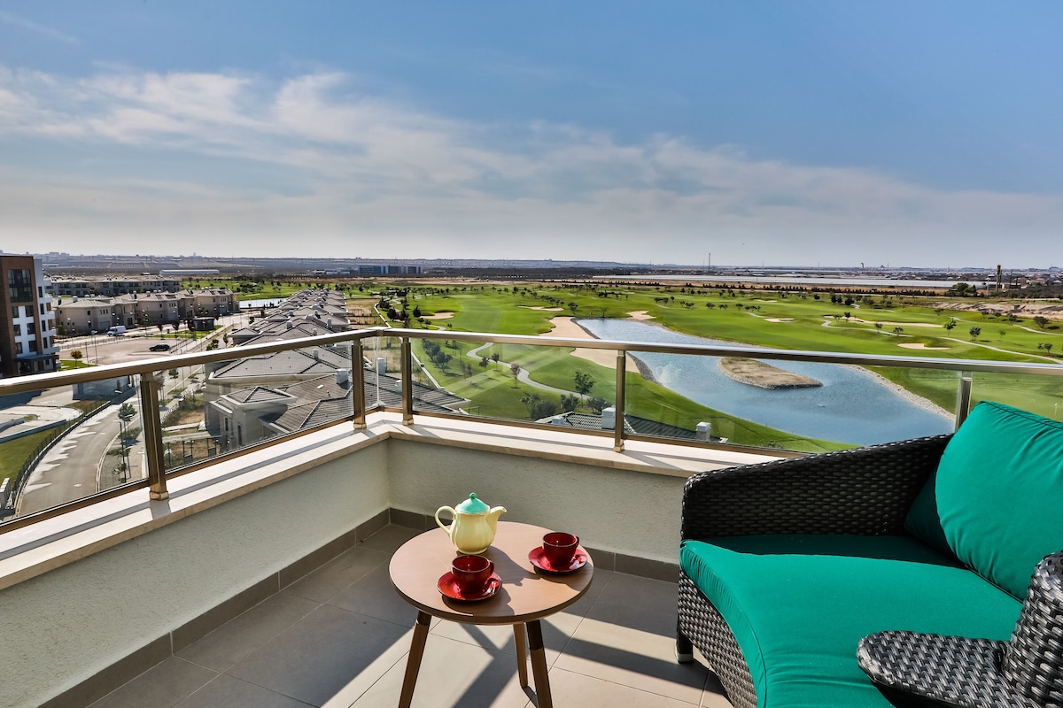 A balcony with a view of a golf course, a wicker sofa, and a table with a teapot and cups.