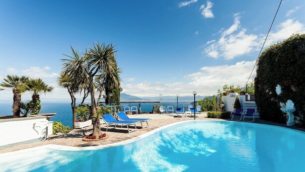 A pool area with lounge chairs and palm trees, overlooking a body of water and distant mountains. - Villa Angela with private pool and sea view (Priora)