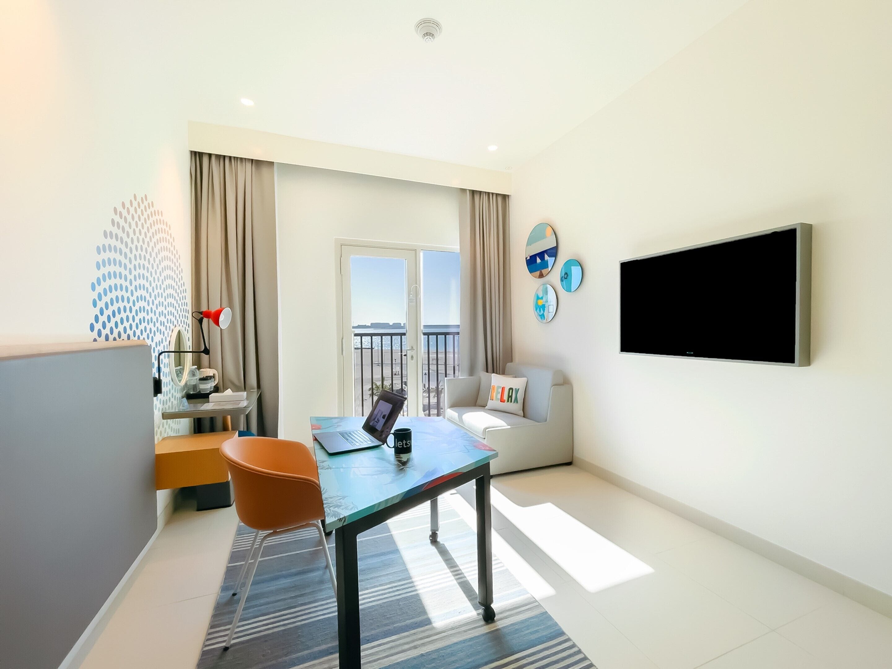 Ein modernes Hotelzimmer mit Balkon, Schreibtisch mit Laptop, Fernseher und Sitzecke mit Sofa und Sessel.