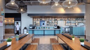 A modern cafe with wooden tables, hanging white pendant lights, and a blue counter with various items. - Rove La Mer Beach (Dubai)