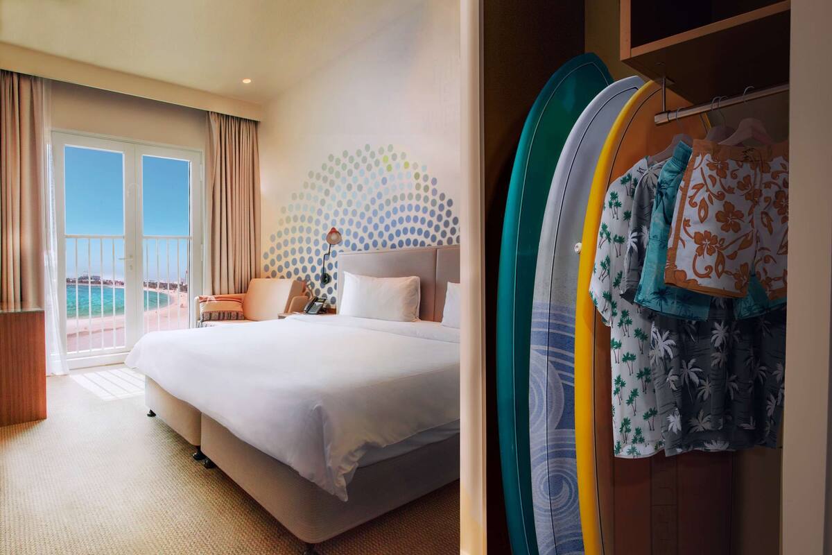 A hotel room with a bed, a desk, a chair, a view of the beach, and a wardrobe with clothes.
