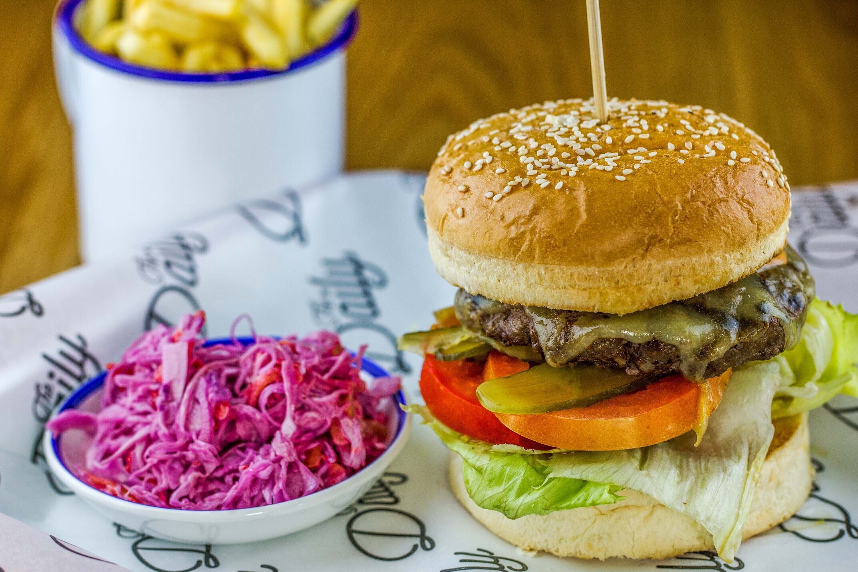 Una hamburguesa con pan de sésamo, lechuga, tomate, pepinillos y una hamburguesa de carne, acompañada de una ensalada de col y papas fritas.
