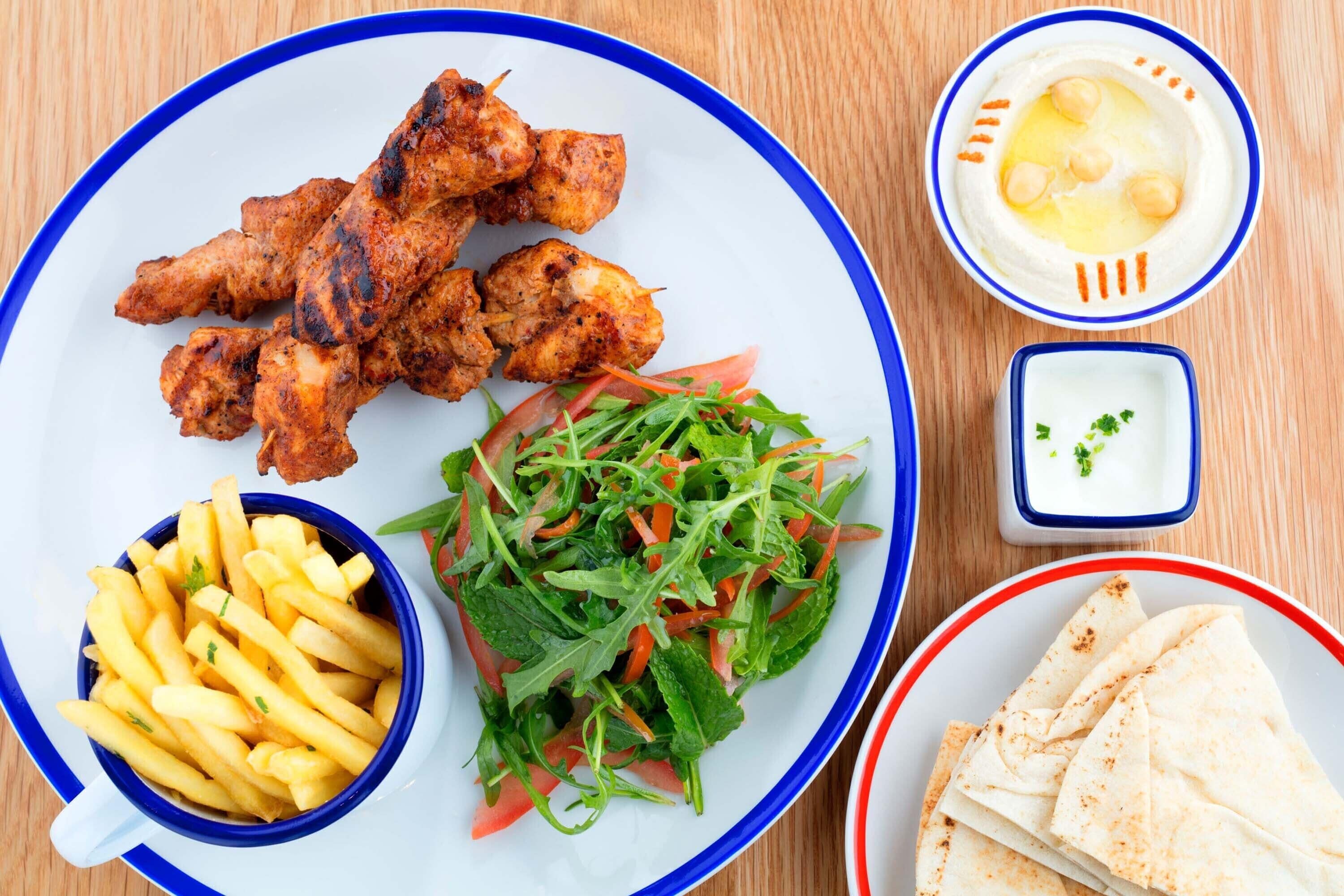A plate with grilled chicken, French fries, a fresh salad, hummus, and pita bread.