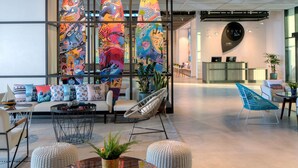 A modern hotel lobby with a seating area, colorful surfboard art, and a reception desk. - Rove La Mer Beach (Dubai)