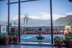 Una persona sentada sobre una colchoneta de yoga con los brazos extendidos, rodeada de plantas en macetas, en un balcón con vistas a montañas nevadas.