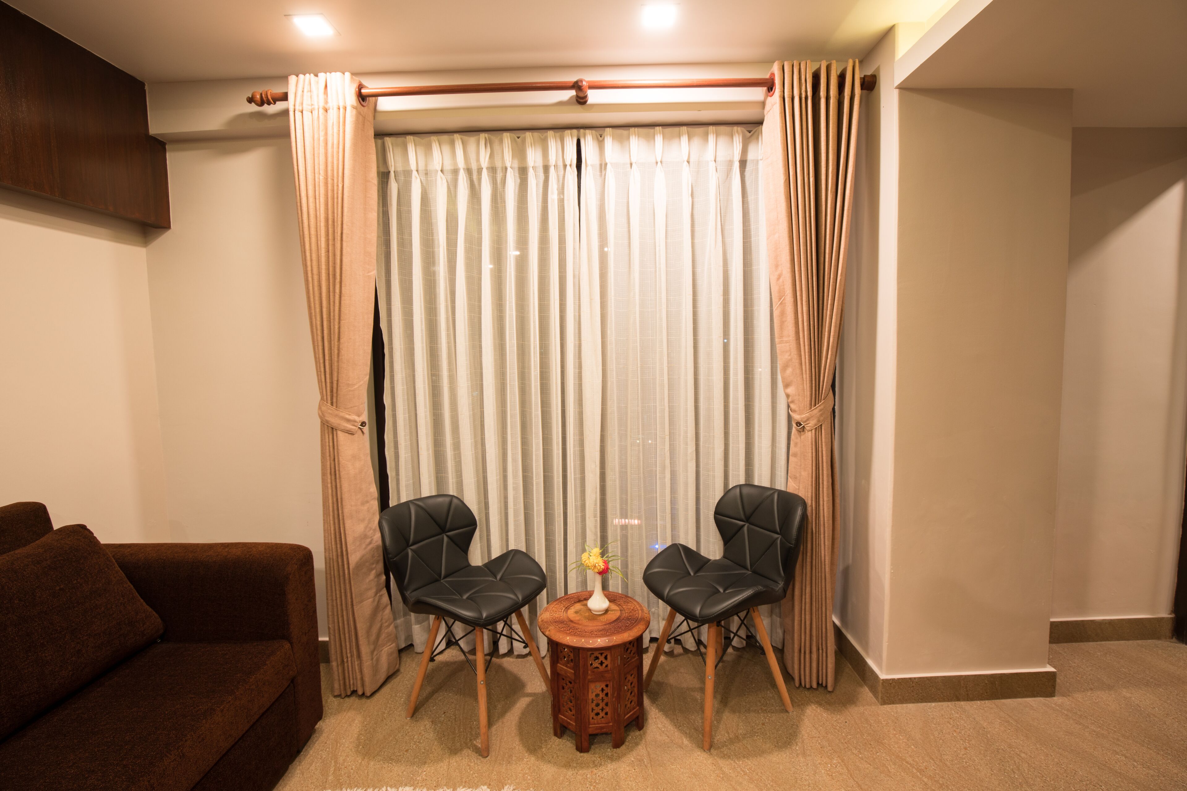 A living room with a brown sofa, two black chairs, a small wooden table with a vase, and a large window with sheer curtains.