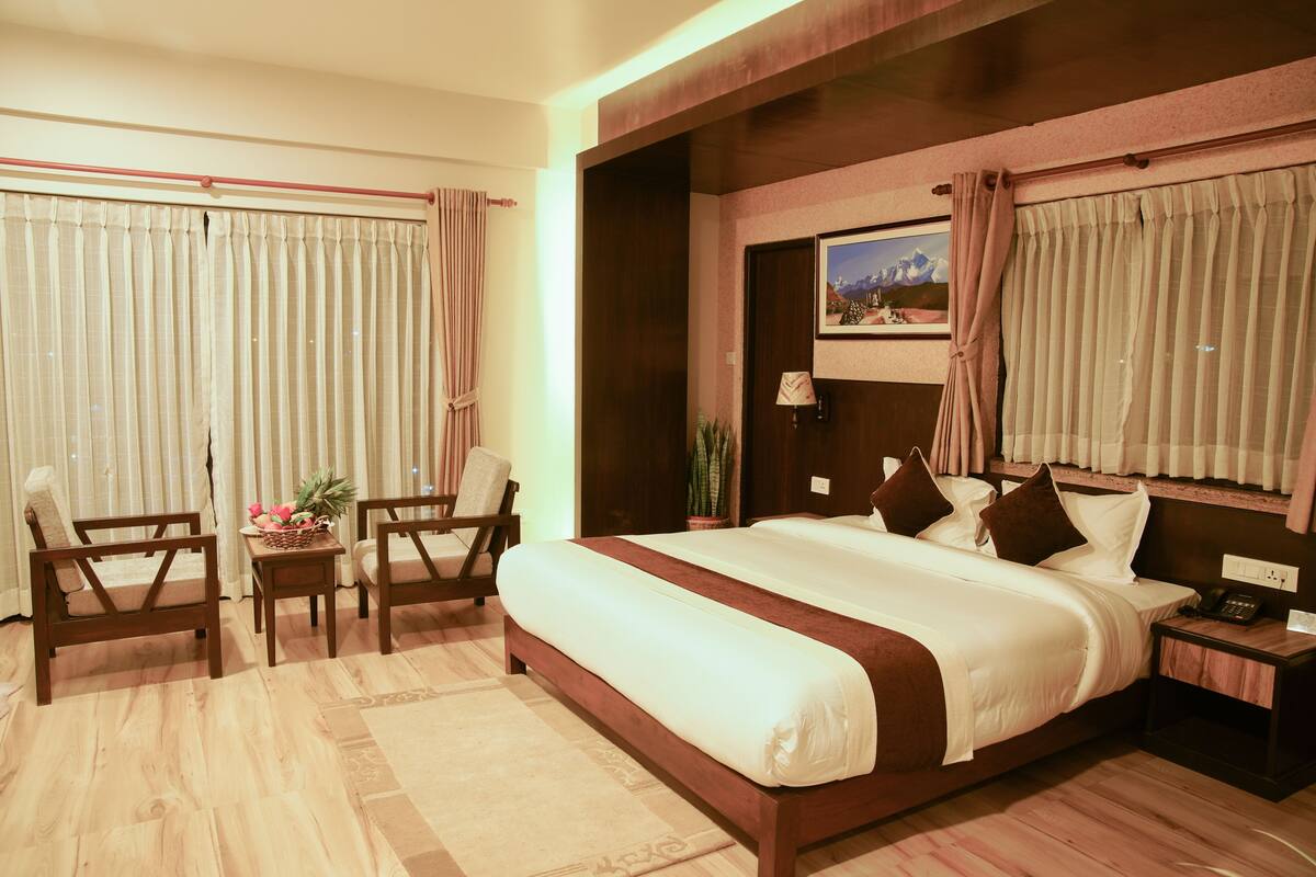 Una habitación de hotel con una cama grande, dos sillas de madera, una mesita con una cesta y una mesita de noche con una lámpara.