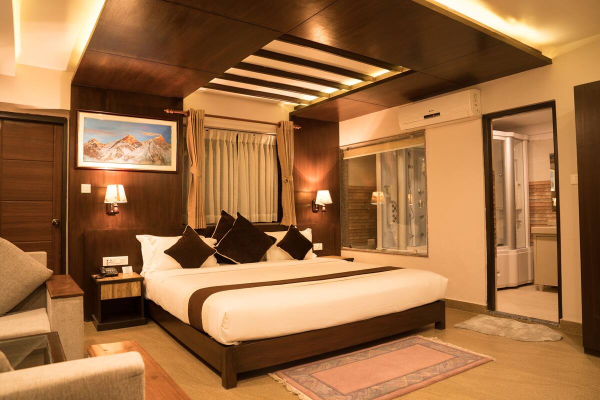 Habitación de hotel con una cama grande, una mesita de noche, una lámpara, un cuadro y un baño visible a través de una puerta abierta.