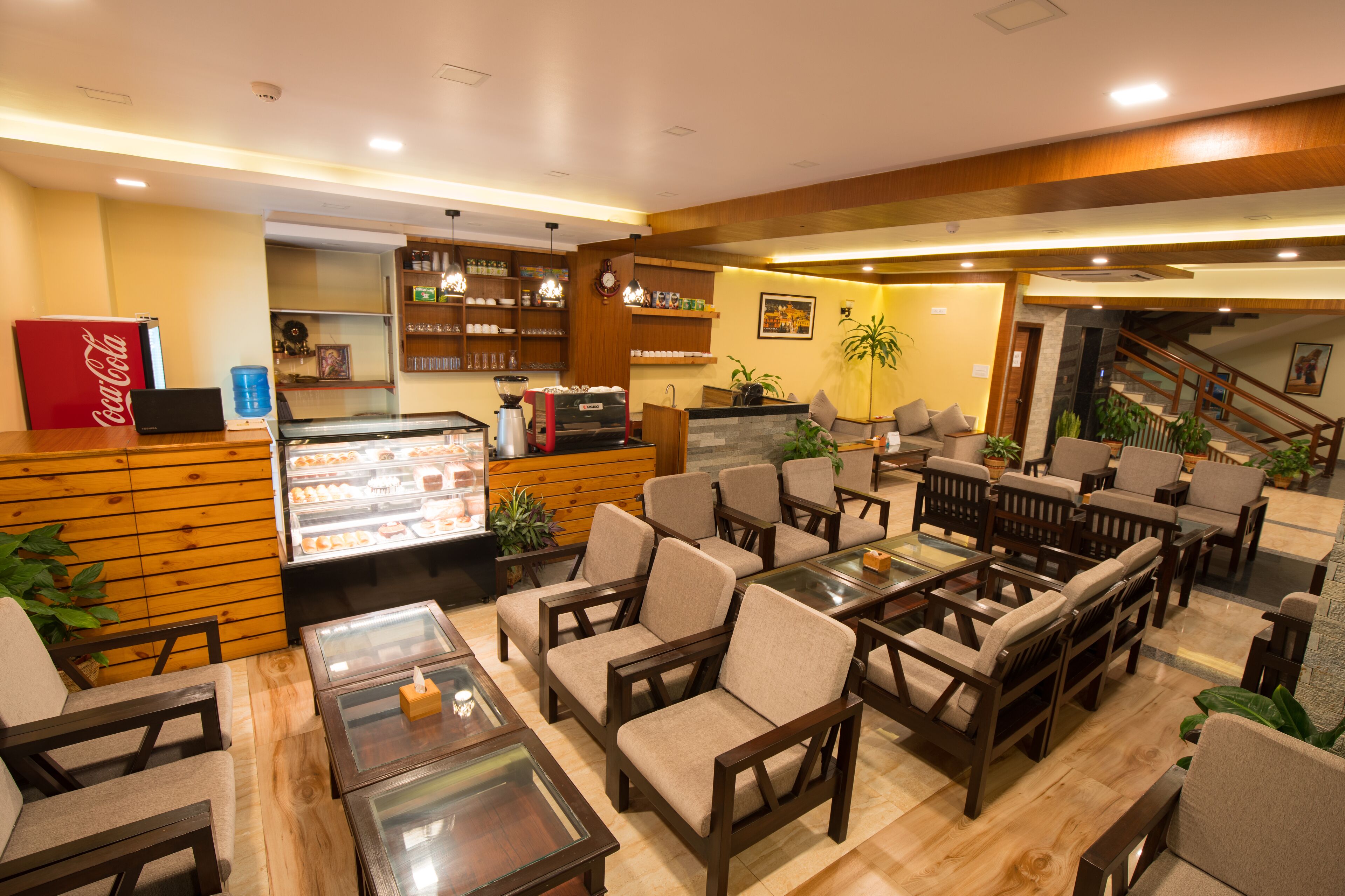 A hotel lobby with wooden paneling, a Coca-Cola refrigerator, a coffee machine, and multiple seating areas with tables and chairs.