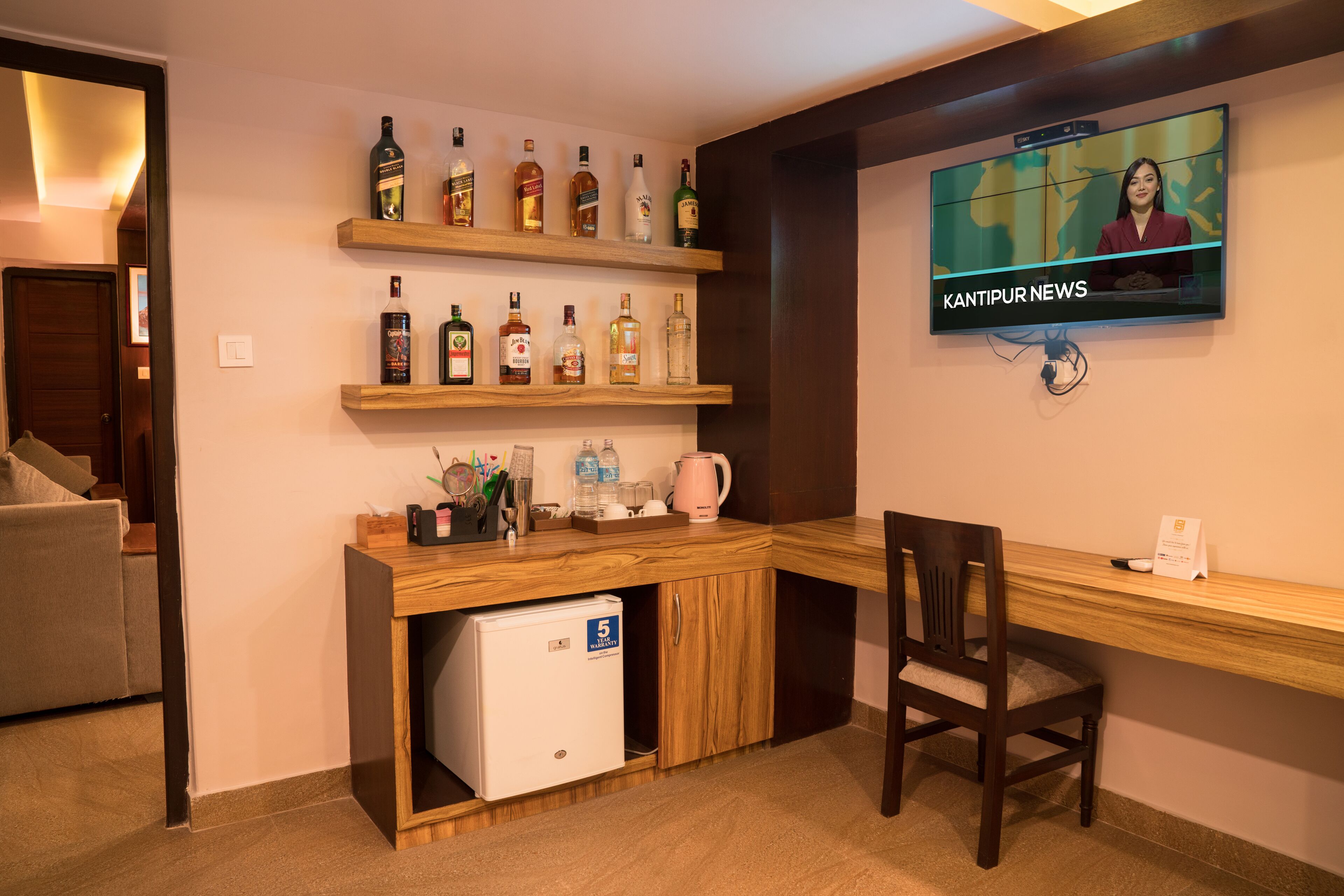 A room with a wooden desk, a television displaying 'KANTIPUR NEWS', shelves with various bottles, and a small refrigerator.