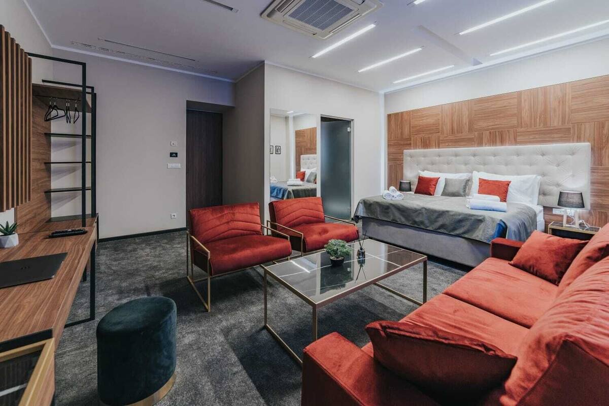 A modern hotel room with a large bed, red upholstered chairs, a glass coffee table, and a wooden accent wall.