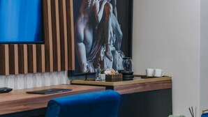 Ein modernes Hotelzimmer mit Schreibtisch aus Holz, blauem Sessel, großem Gemälde an der Wand und einer Kaffeestation.