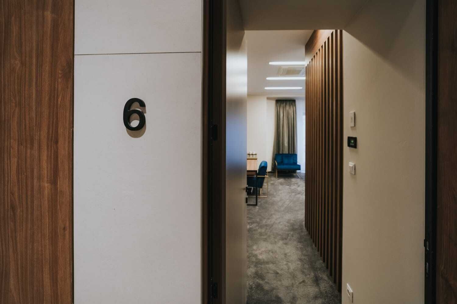 Un pasillo con una puerta marcada con el número 6, que da a una habitación con un sofá azul y un escritorio.