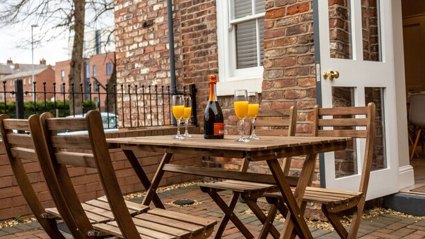 Een houten buitentafel, gedekt met stoelen, een fles en glazen sinaasappelsap.