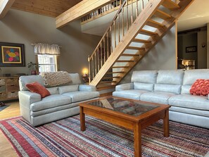 A living room with two sofas, a glass coffee table, a wooden staircase, and a view of the kitchen.